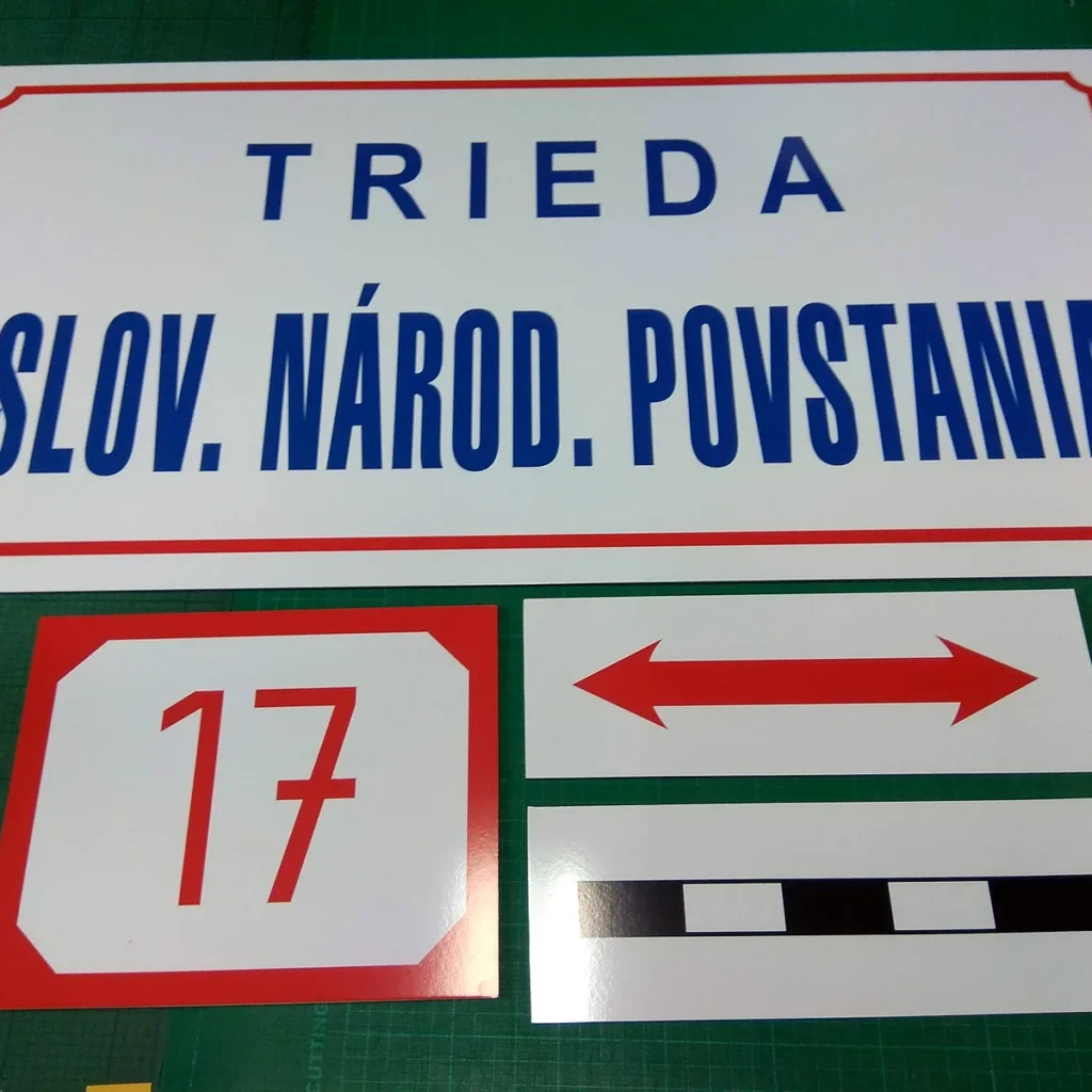 Biela tabuľa s červeným rámikom a modrým nápisom názvu ulice, vedľa nej červené číslo 17 a obojstranná šípka