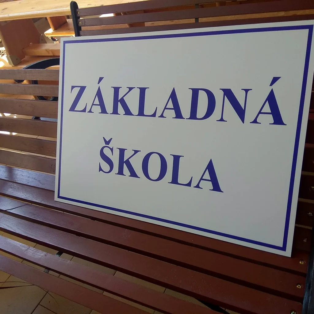 Biela tabuľa s modrým nápisom Základná škola a modrým rámom oprená o drevenú lavičku