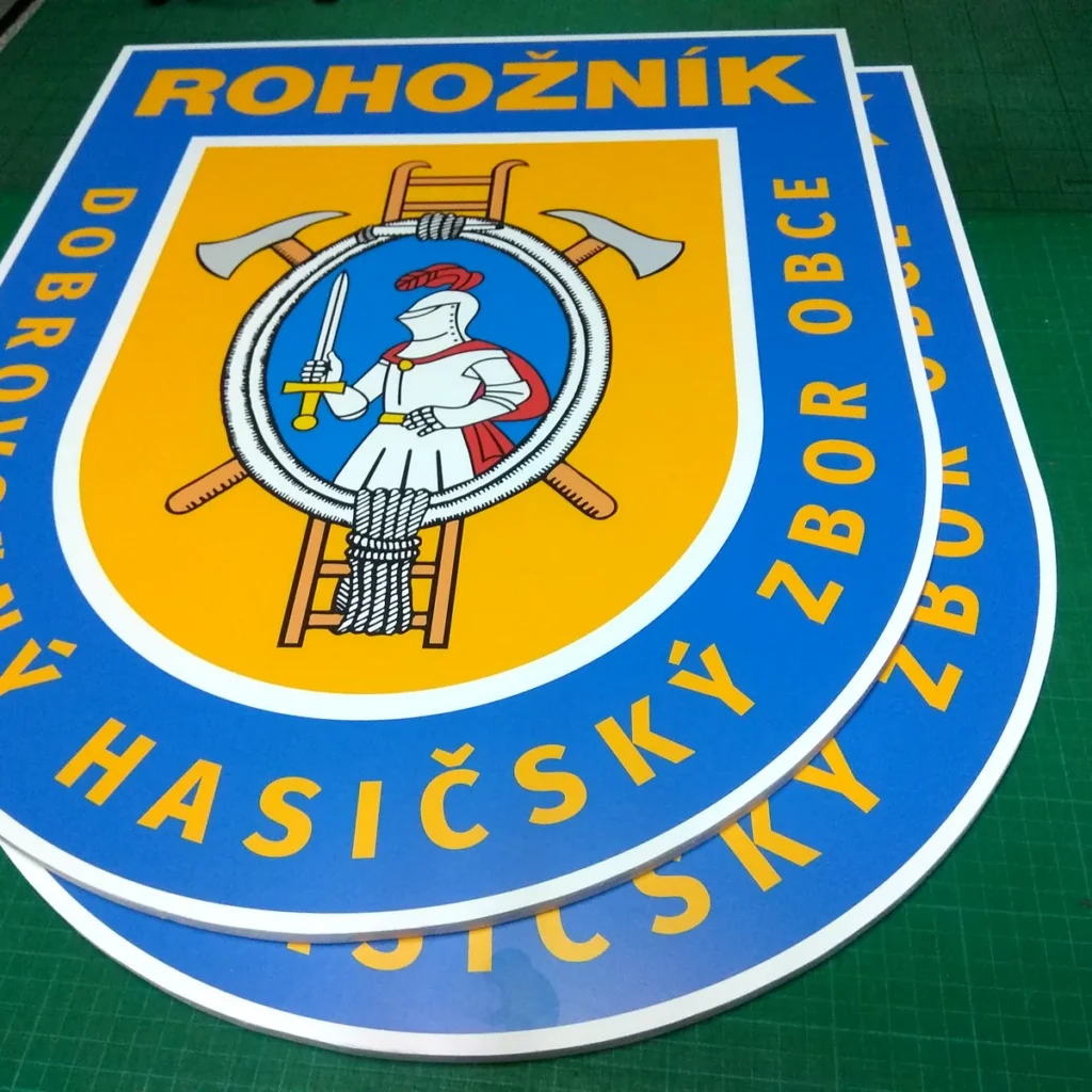Dva farebné erby hasičského zboru vyrezané po obryse z hrubej bielej dosky na zelenej podložke stola